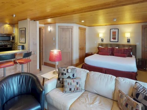 A cozy studio apartment with a bed, sofa, armchair, kitchen area, and a lamp on an end table. Warm wooden ceiling and modern decor adorn the space.