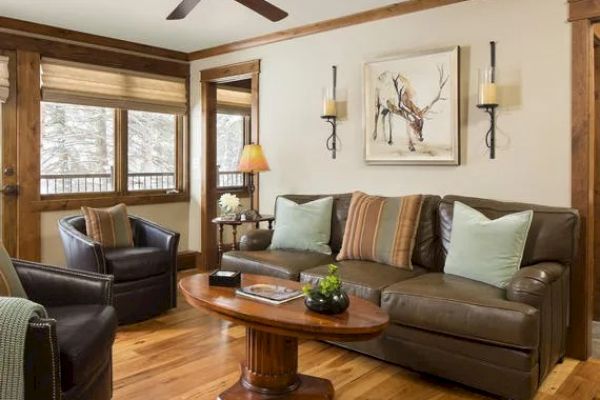 A cozy living room with a leather sofa, armchairs, wooden coffee table, wall art of a deer, and windows letting in natural light.