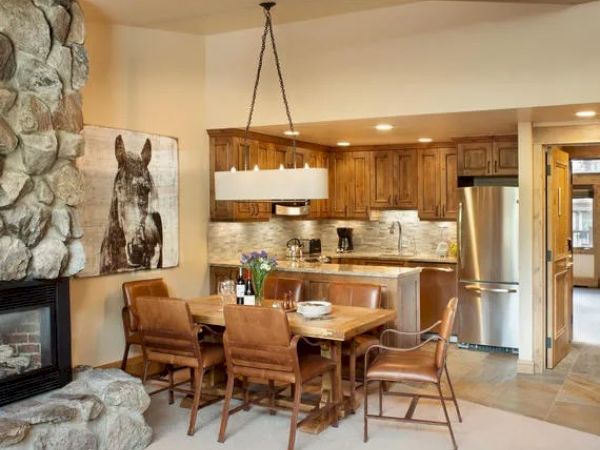 A cozy dining area with a wooden table, leather chairs, a stone fireplace, and a kitchen with wooden cabinets and stainless steel appliances.