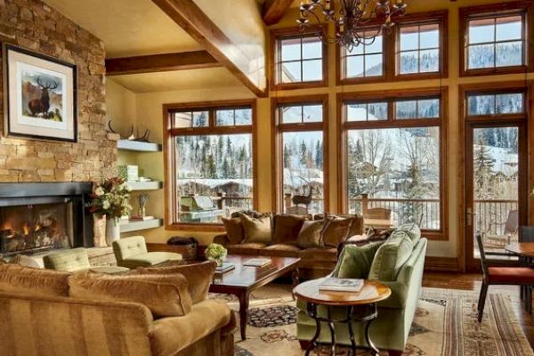 A cozy living room with large windows, mountain views, a stone fireplace, and comfortable seating in a cabin-style interior.