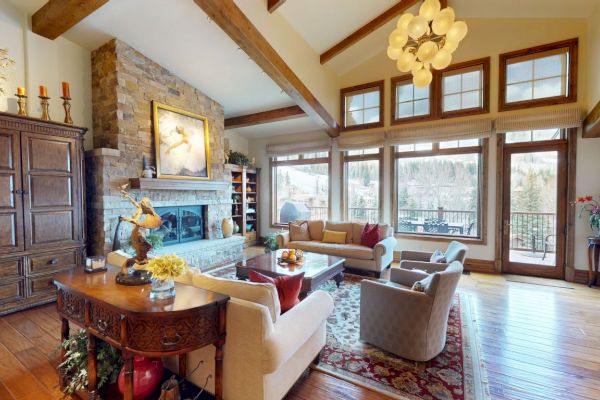 A cozy living room with wooden beams, large windows, stone fireplace, elegant furniture, and outdoor view.