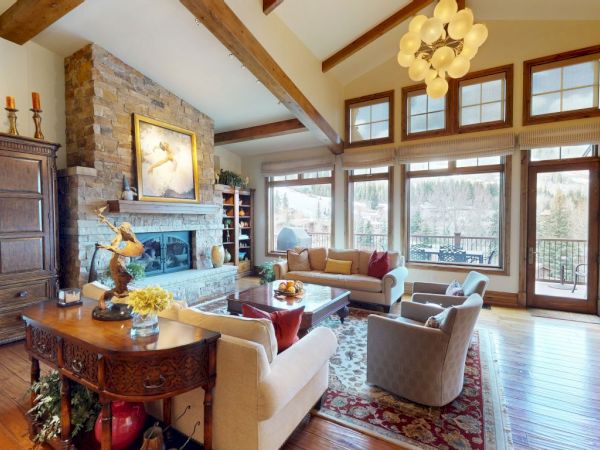 A cozy living room with wooden beams, large windows, stone fireplace, elegant furniture, and outdoor view.