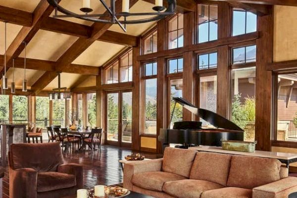 A spacious living room features large windows, wooden beams, a grand piano, a dining area, and cozy seating with a coffee table.