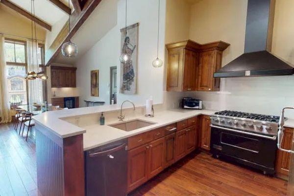 A modern kitchen with wooden cabinets, a large island, stainless steel appliances, and pendant lights. It features high ceilings and hardwood floors.