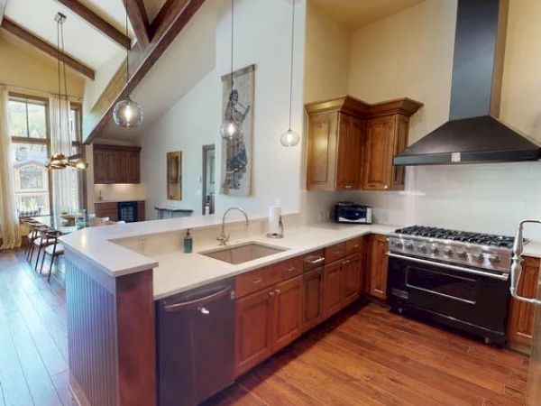A modern kitchen with wooden cabinets, a large island, stainless steel appliances, and pendant lights. It features high ceilings and hardwood floors.