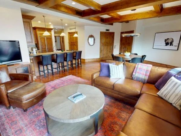 A cozy living area with a large brown leather sectional sofa, armchair, round coffee table, kitchen with barstools, and modern decor, including a wall picture.