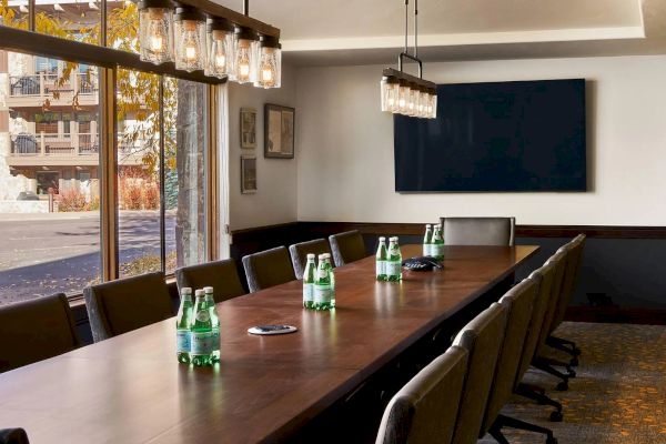 A conference room with a long table, chairs, water bottles, a large screen on the wall, and large windows allowing natural light in.