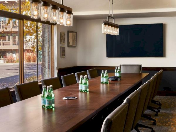 A conference room with a long table, chairs, water bottles, a large screen on the wall, and large windows allowing natural light in.