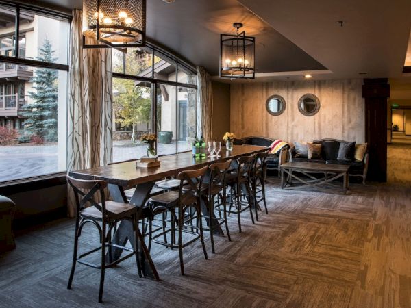 A cozy, modern room featuring a long wooden table with chairs, a couch, decorative lighting, and large windows with views of trees and buildings outside.