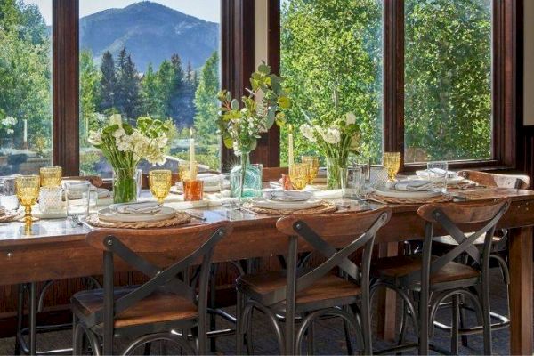 A dining table is set with floral arrangements, colorful glassware, and plates, positioned next to large windows showcasing a scenic mountain view.