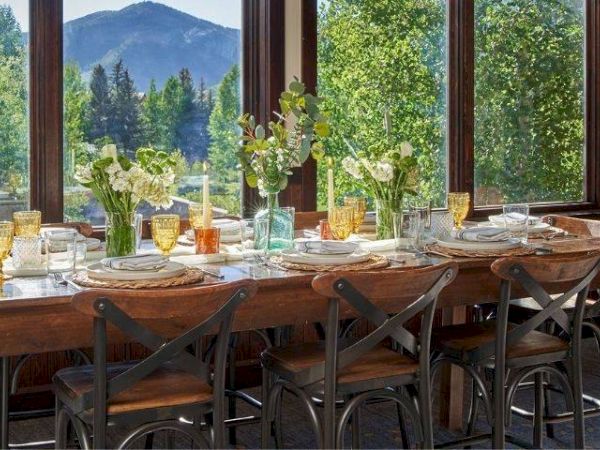 A dining table is set with floral arrangements, colorful glassware, and plates, positioned next to large windows showcasing a scenic mountain view.