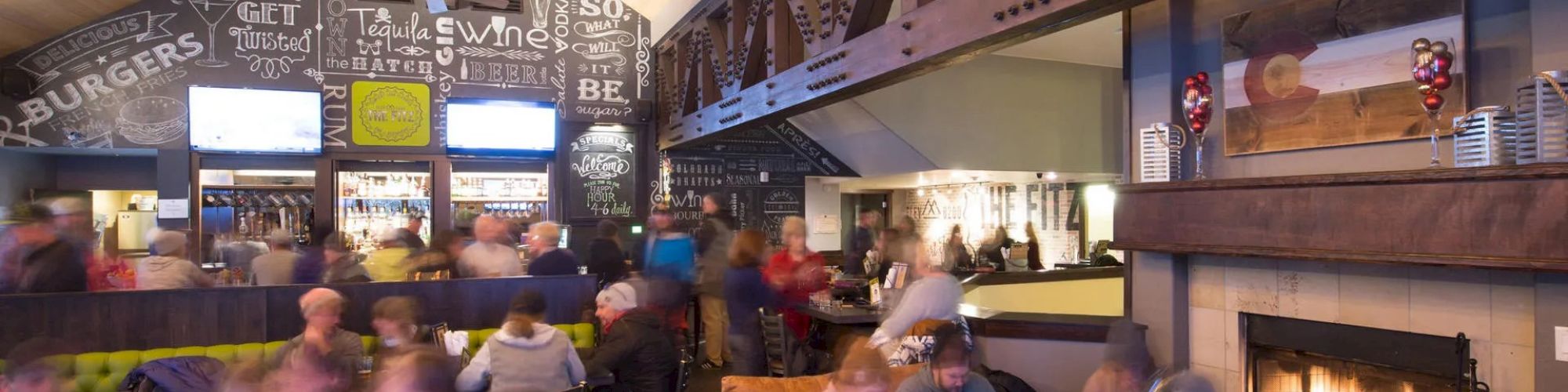 A busy, modern indoor brewery or bar with people seated and socializing, a large menu chalkboard, and a cozy fireplace on the right.