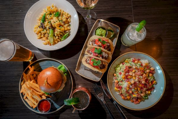 A table with a burger and fries, tacos, pasta, and a salad, accompanied by cocktails and a beer.