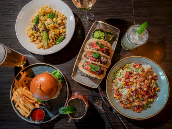 A table with a burger and fries, tacos, pasta, and a salad, accompanied by cocktails and a beer.