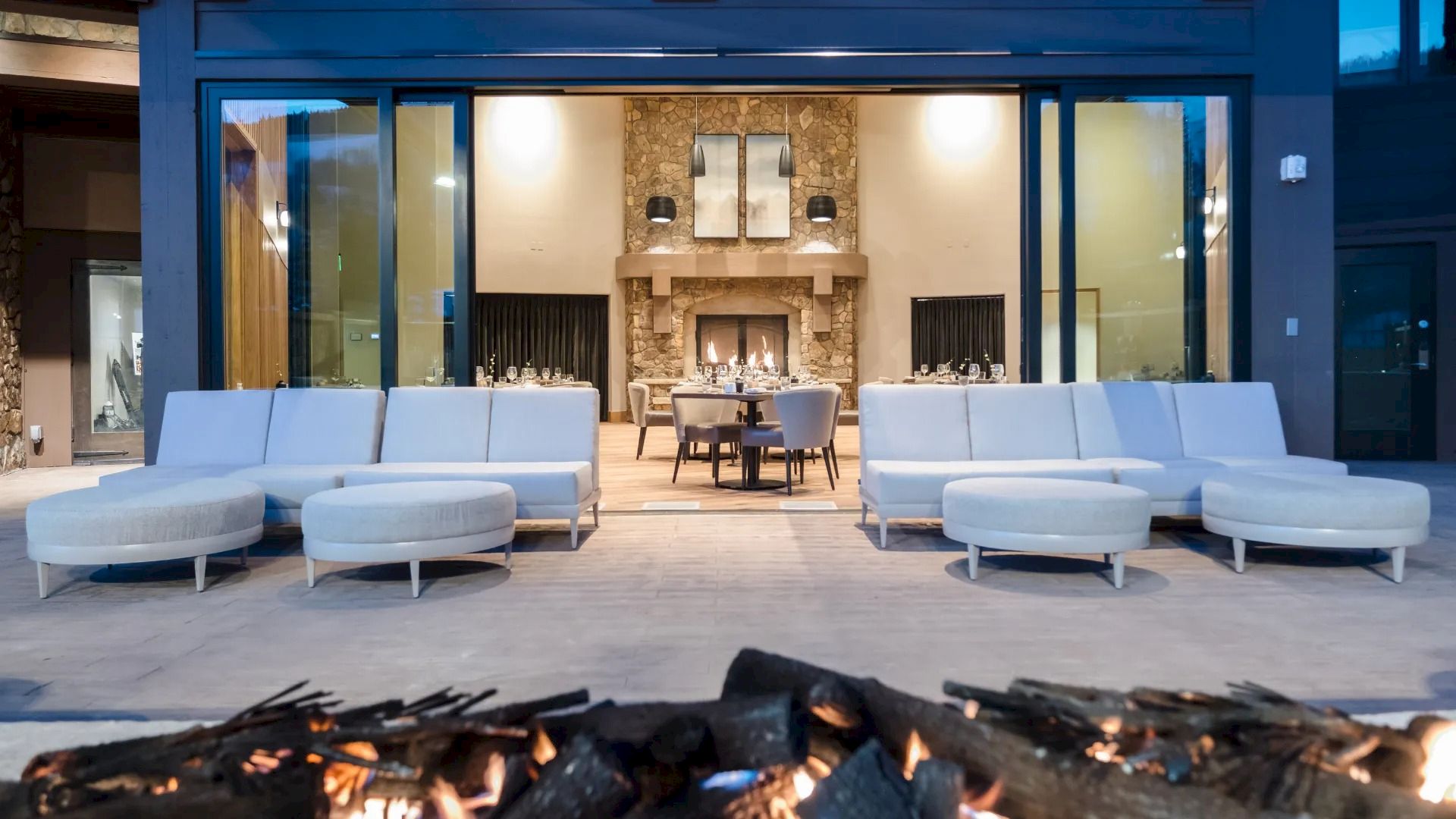 Modern patio with white seating, fire pit in the foreground, and a view into an elegant indoor dining area with a stone fireplace, ending the sentence.