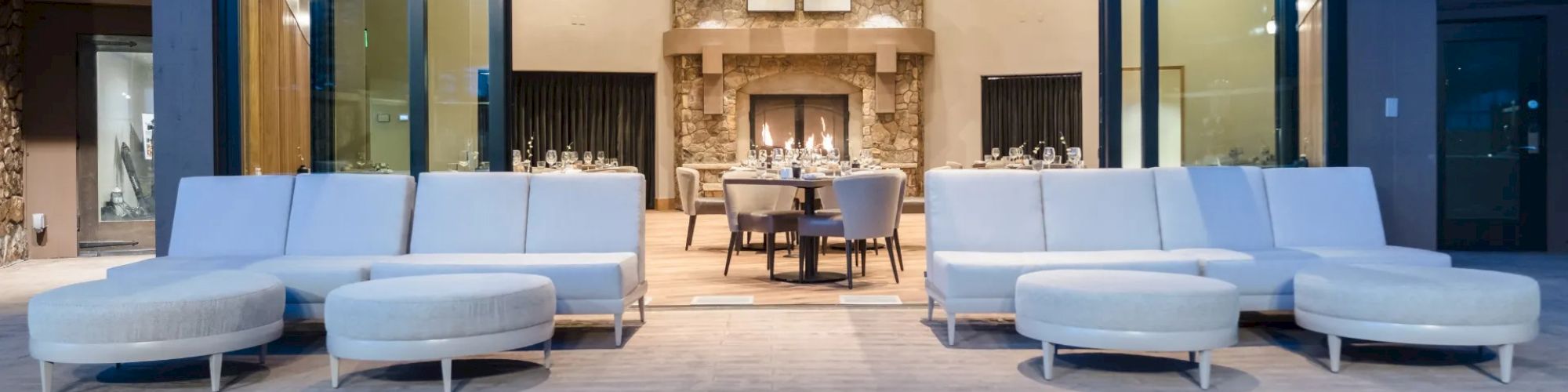 Modern patio with white seating, fire pit in the foreground, and a view into an elegant indoor dining area with a stone fireplace, ending the sentence.