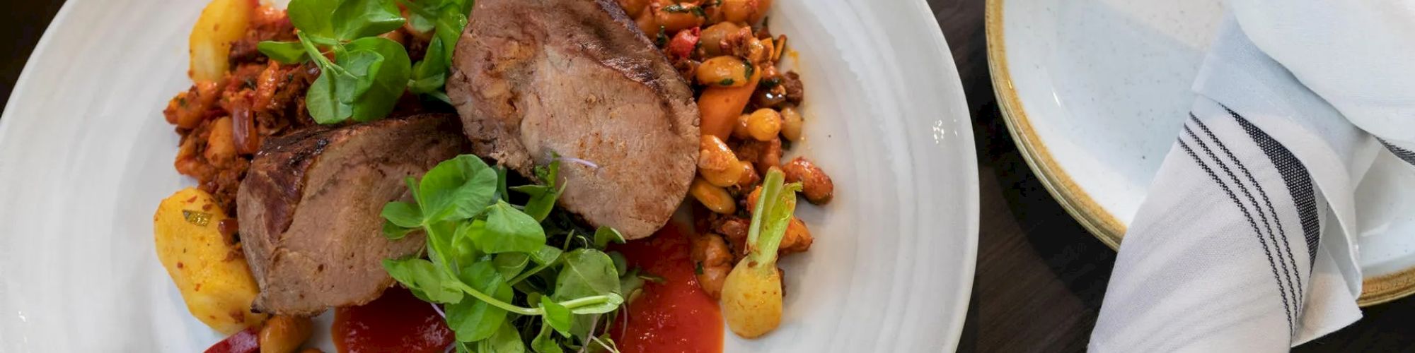 A plated dish featuring two slices of meat atop grains and legumes with greens, served with a tomato-based sauce, and a napkin on the side.