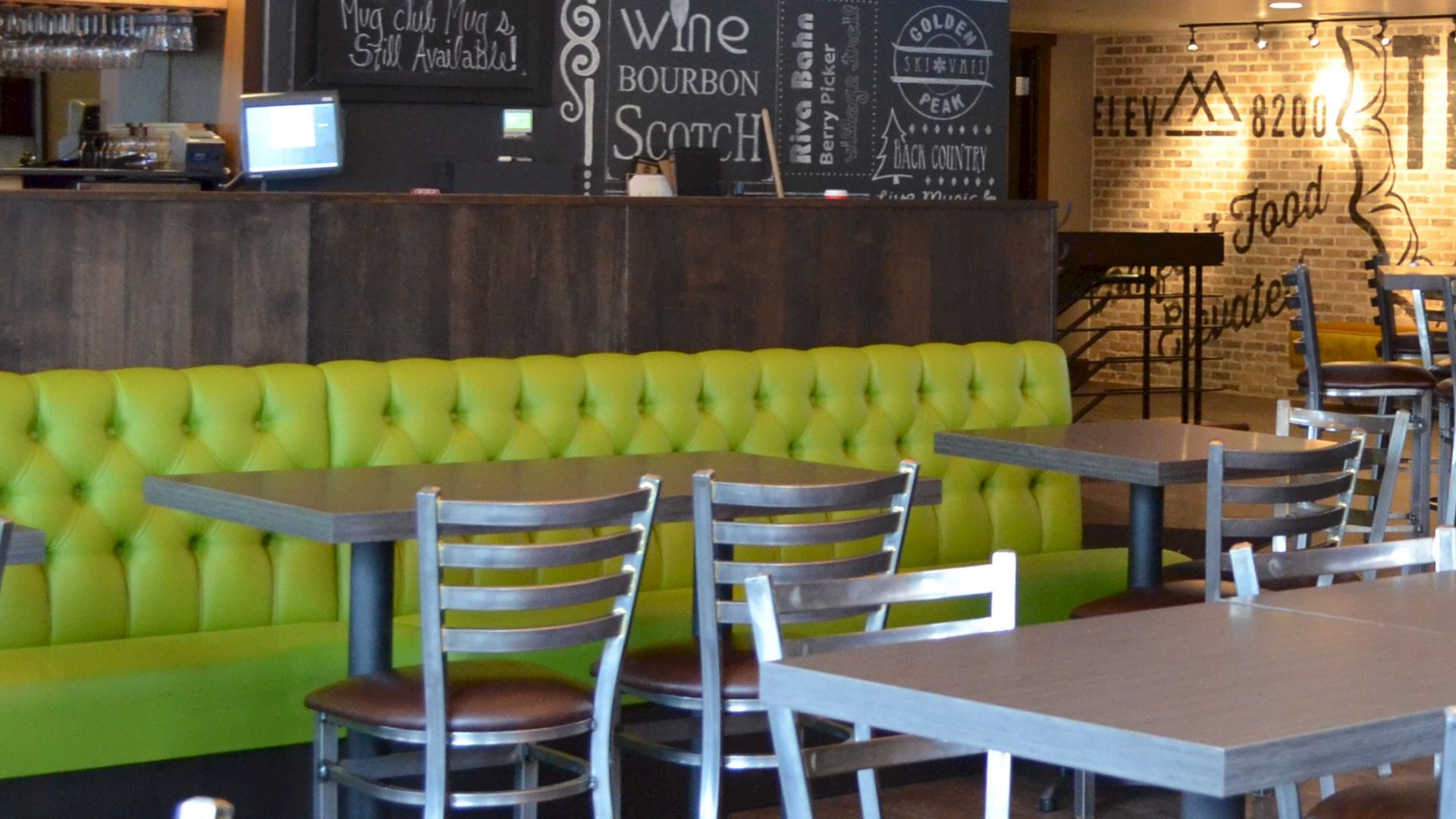A restaurant interior with green seating, wooden tables, metal chairs, and a chalkboard menu on the wall.