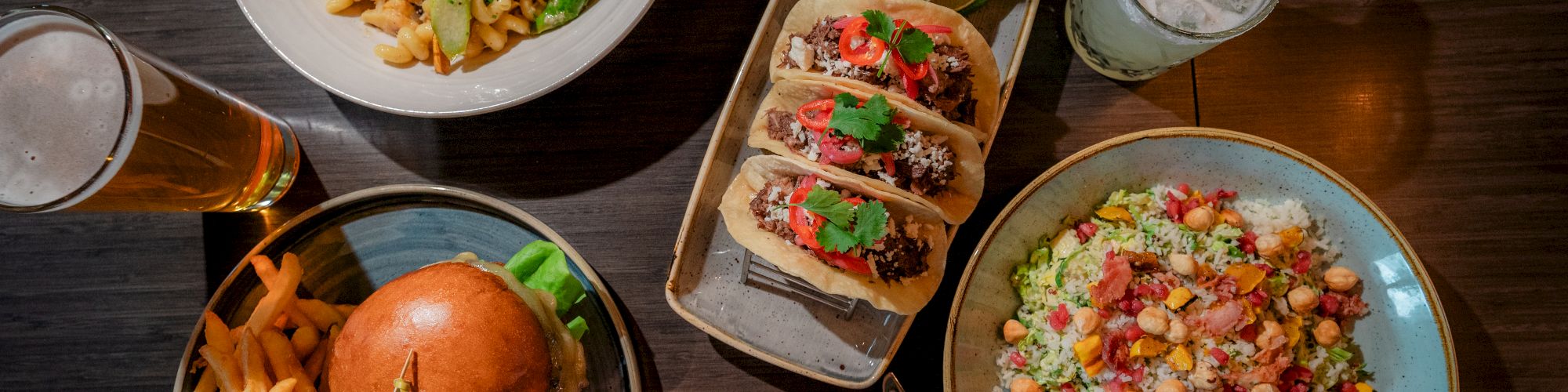 An array of dishes: pasta, tacos, burger with fries, salad, and drinks including cocktails and beer on a wooden table.