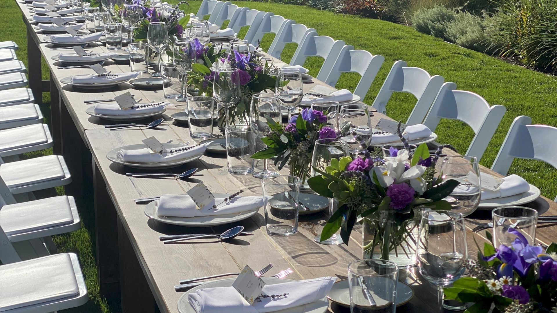 An outdoor dining setup with a long table, white chairs, place settings, and purple floral centerpieces on a grassy area under the sun.