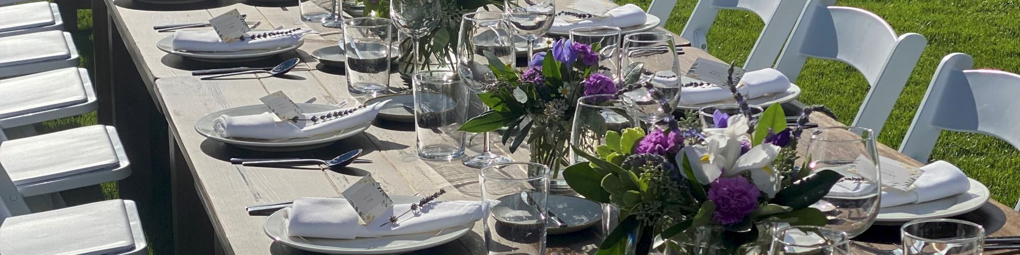 An outdoor dining setup with a long table, white chairs, place settings, and purple floral centerpieces on a grassy area under the sun.