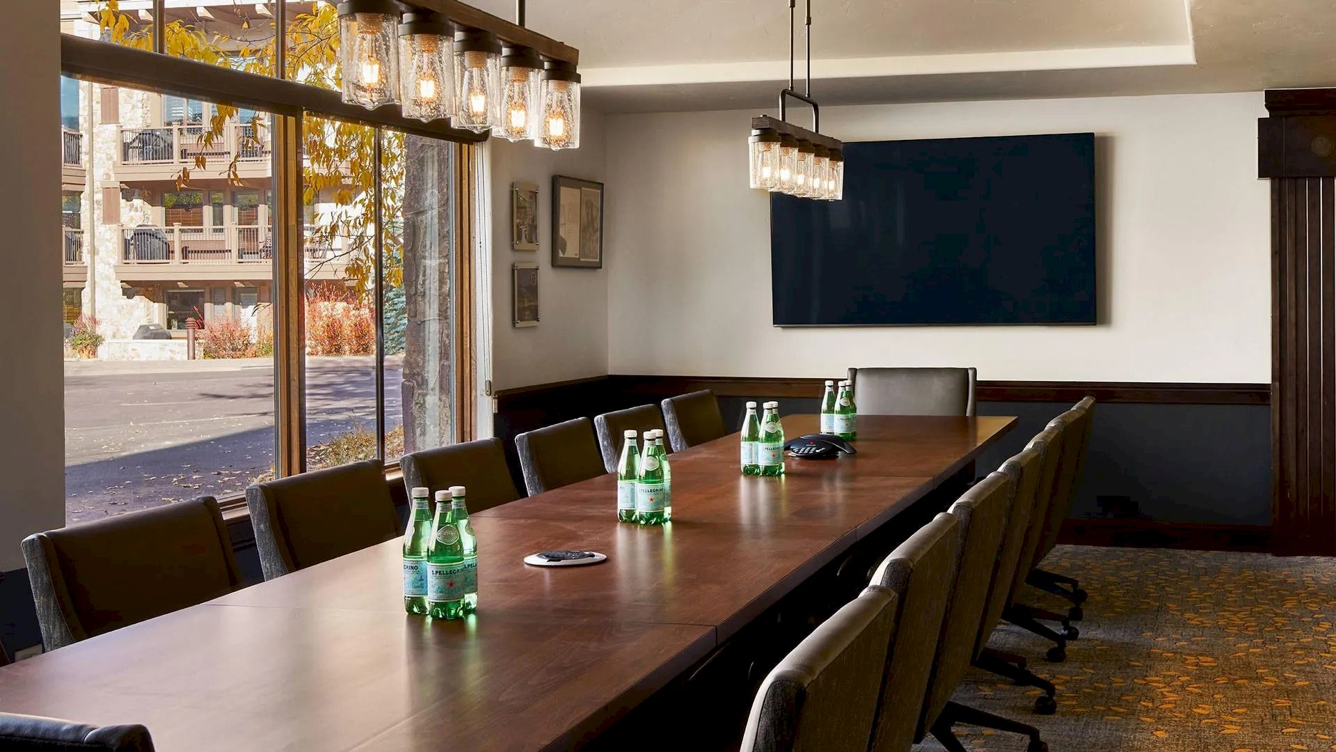 A modern conference room with a long wooden table, chairs, water bottles, wall-mounted screen, large windows, and hanging lights.