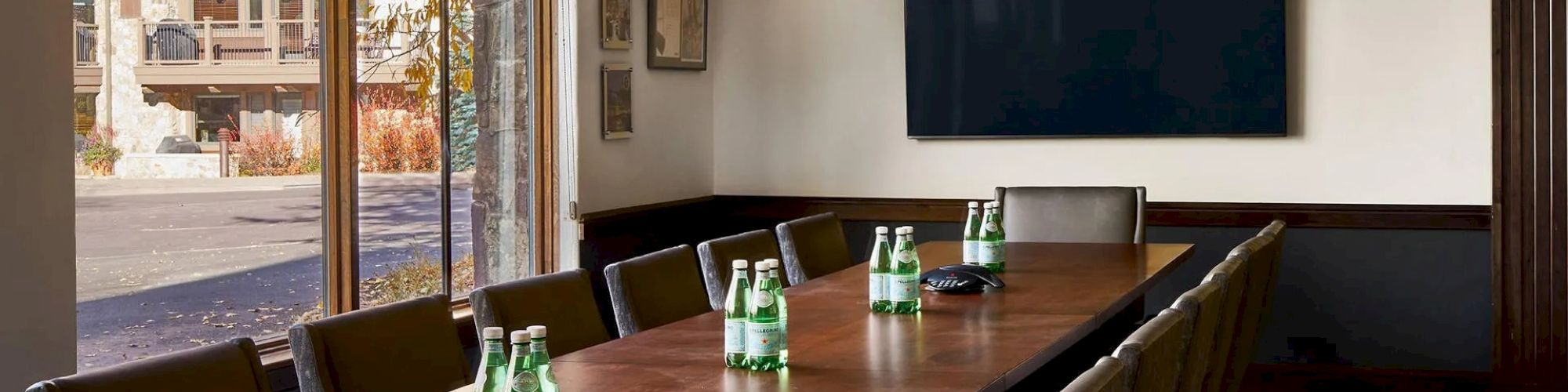 A modern conference room with a long wooden table, chairs, water bottles, wall-mounted screen, large windows, and hanging lights.