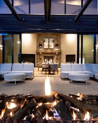 The image shows an outdoor seating area with a modern fireplace, white couches, and a view into an elegant interior with more seating.