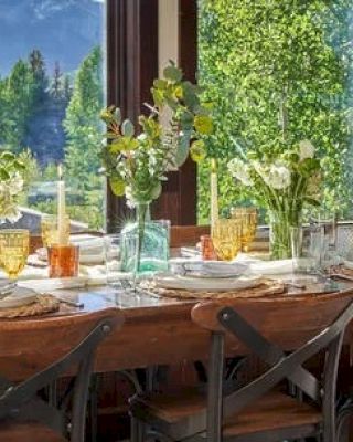 A beautifully set dining table is arranged with flowers, glassware, and tableware, situated in a room with large windows overlooking a lush, green landscape.