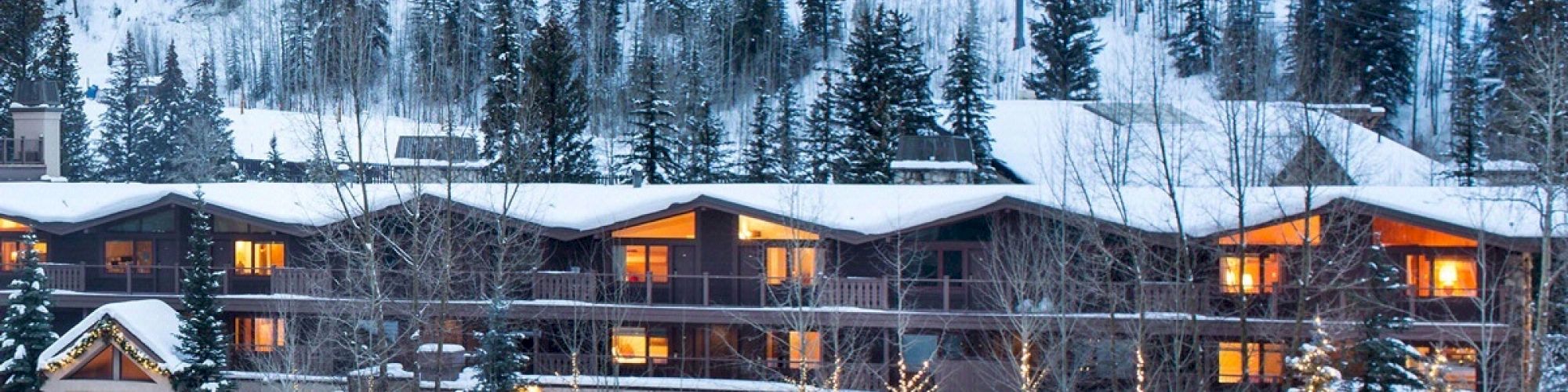 A cozy, snow-covered lodge nestled in a wintery pine forest, with warm lights glowing from the windows, ski lift and slopes in the background.