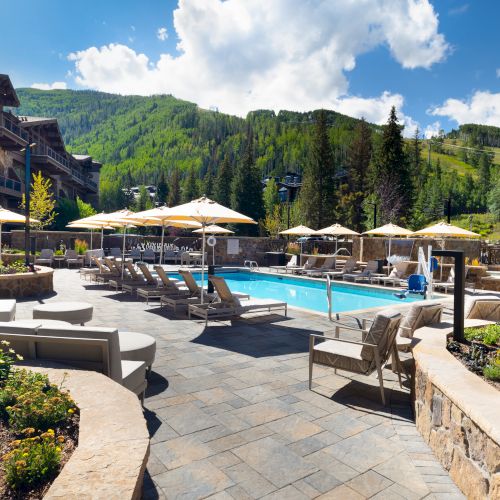 An outdoor pool area with lounge chairs and umbrellas, surrounded by mountains and greenery under a blue sky with clouds.