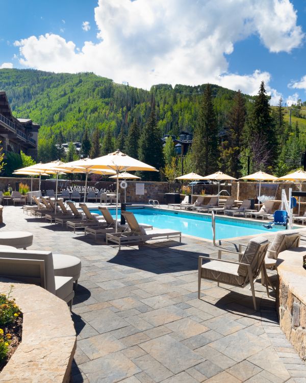 An outdoor pool area with lounge chairs and umbrellas, surrounded by mountains and greenery under a blue sky with clouds.