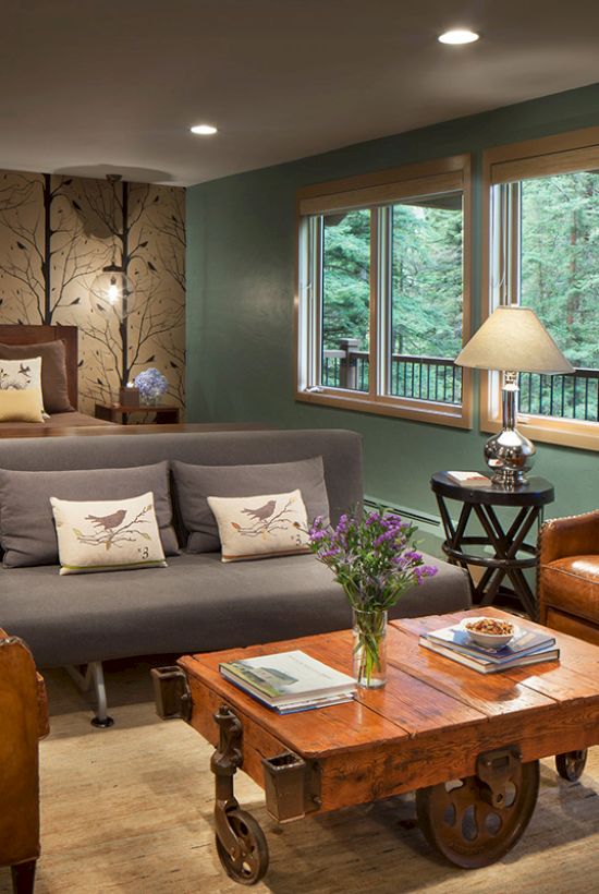 A cozy room with a bed, sofa, two leather chairs, a coffee table, table lamp, and large windows offering a forest view.