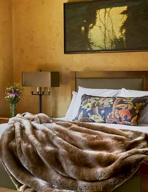 A cozy bedroom with a large window, bed with a fur blanket, floral cushion, bedside table with flowers, and a wall painting above the bed.