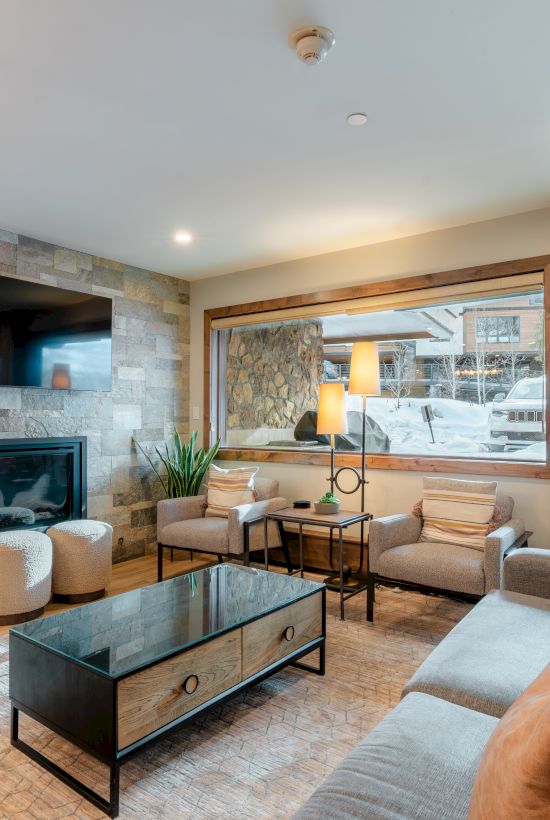 A cozy living room with a stone fireplace, wall-mounted TV, large windows, modern furniture, and a scenic snowy view outside.