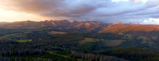 The image shows a breathtaking landscape with mountains under a cloudy sky, dense forests, and patches of open fields illuminated by sunlight.