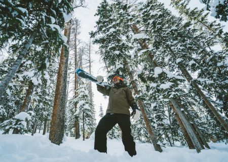 A person stands in a snowy forest holding a snowboard, surrounded by tall trees covered in snow, wearing winter sports gear.