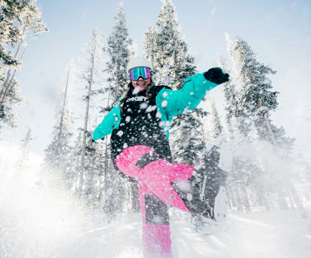 A person snowboarding down a snowy slope, kicking up powder, with snow-covered trees in the background under a clear blue sky.