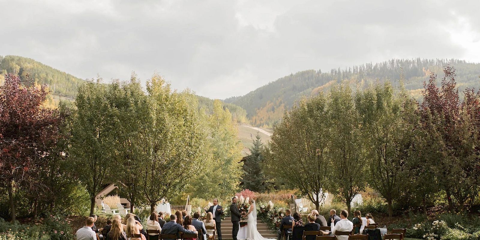 An outdoor wedding ceremony is taking place in a scenic, nature-filled location with trees and mountains in the background, attended by several guests.