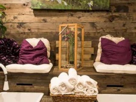 A cozy spa setup with wooden walls, chairs with purple cushions, a basket of white rolled towels, and a framed flower art piece on the wall.