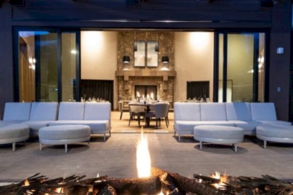 The image shows a cozy outdoor patio with a fire pit, white seating, and a view into a warmly lit interior with a stone fireplace.