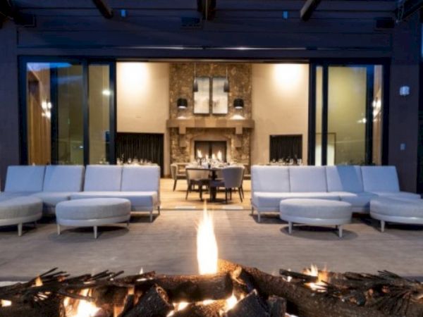 The image shows a cozy outdoor patio with a fire pit, white seating, and a view into a warmly lit interior with a stone fireplace.
