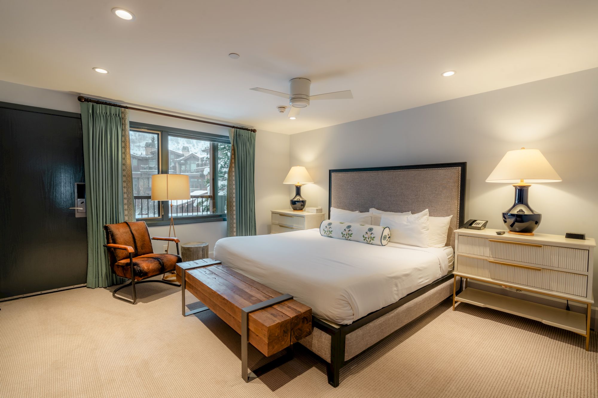 A modern bedroom with a large bed, bedside tables with lamps, a wooden bench, and a window with green curtains.