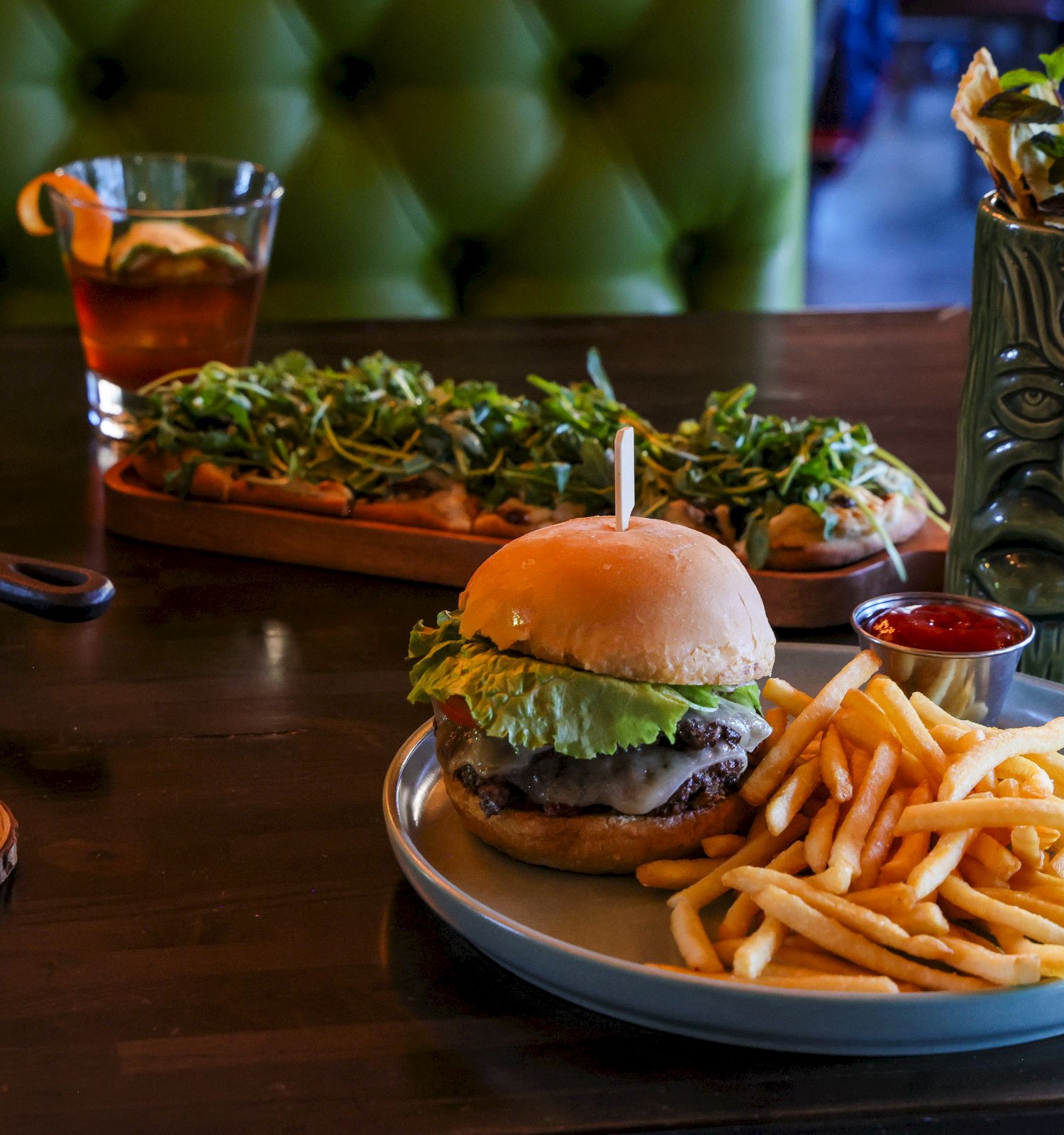 A sizzling burger with lettuce, fries, and a glossy bun sits on a plate, while two drinks and a green herb garnish decorate the wooden table.