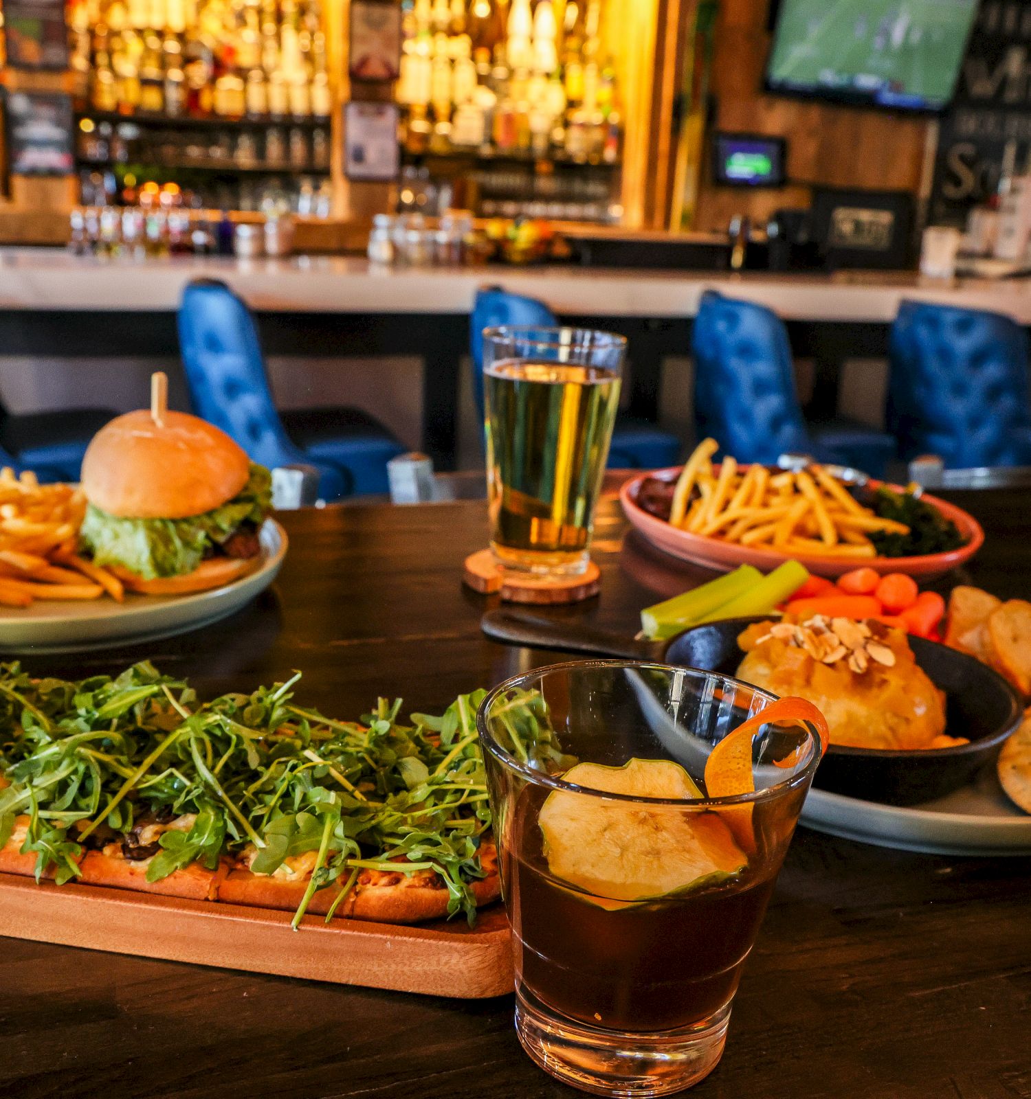 A lively restaurant spread: burgers, fries, wings, greens, and drinks on a dark wooden table with a bar backdrop. Ends with a period.