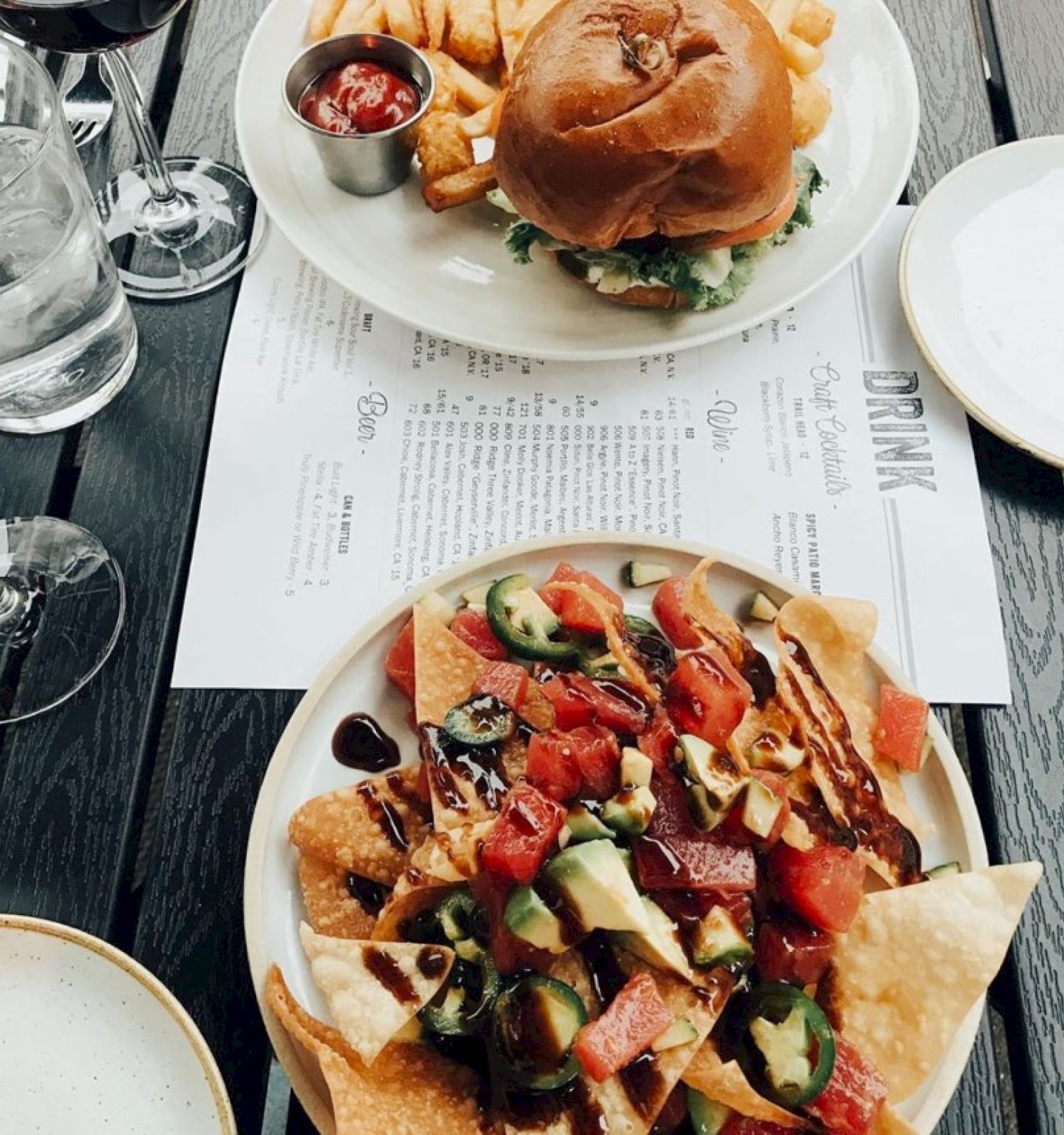 A table with nachos topped with jalapeños and tomatoes, a burger with fries, two glasses of wine, and a menu.