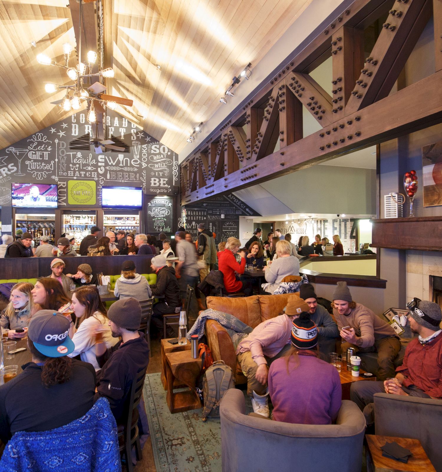 People socializing in a cozy bar with a fireplace, chalkboard art, and rustic beams, creating a lively and inviting atmosphere.