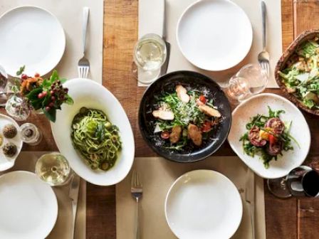 A table set with plates of salad, pasta, and appetizers, along with drinks, cutlery, and flowers on placemats, ready for a meal.