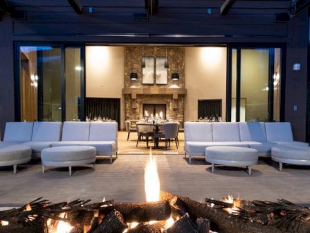 Outdoor patio with modern seating, a fire pit, and a view into a cozy indoor dining area with a fireplace, at dusk.