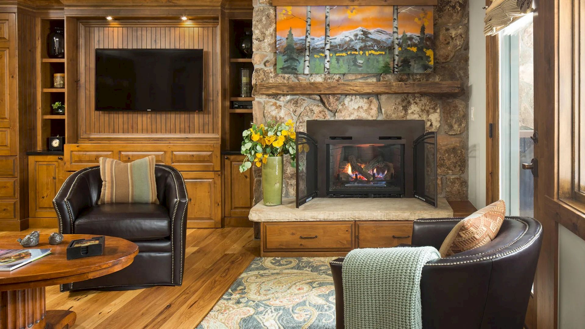 A cozy living room features a stone fireplace, two leather chairs, a wall-mounted TV, a colorful painting, and wooden cabinetry.